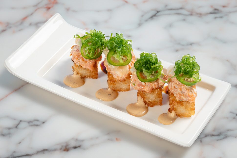 Truluck’s Fort Lauderdale salmon crispy rice featuring four golden crispy rice squares topped with spicy salmon, sliced jalapeños, fresh scallions, and a drizzle of creamy sauce, served on a white rectangular plate over a marble surface.