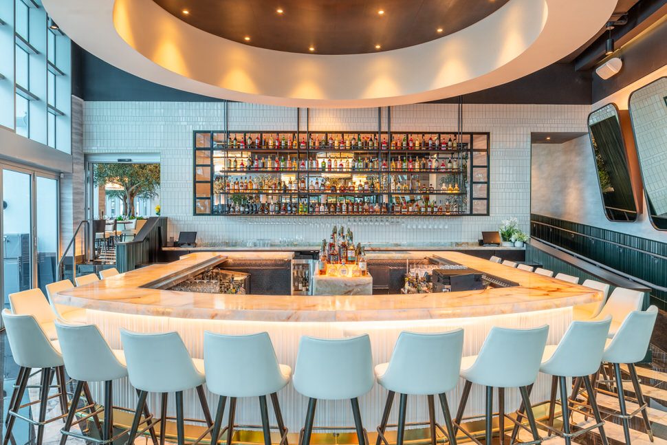 Truluck’s Fort Lauderdale main bar featuring a circular marble countertop with light blue bar stools, an illuminated back bar displaying premium spirits, white tile walls, and a dramatic round ceiling detail with recessed lighting.