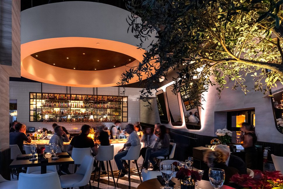 Guests dining and socializing at Truluck’s Fort Lauderdale bar, featuring a glowing circular ceiling detail, illuminated back bar with premium spirits, modern white bar stools, and warm ambient lighting beneath an indoor olive tree.