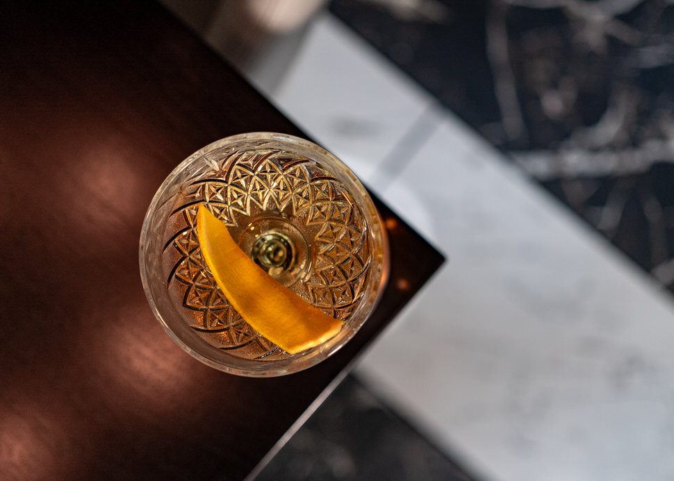 El Tesoro tequila martini viewed from above, served in a patterned crystal glass with an orange peel garnish, resting on a dark wood bar beside a marble surface.