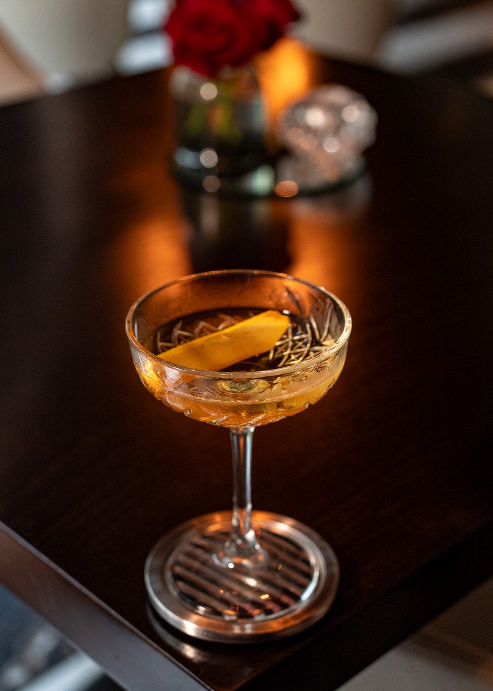 El Tesoro martini served in a cut-crystal coupe glass with a wide lemon peel garnish and rosemary sprigs floating on top. The cocktail rests on a polished dark wood table with a metal coaster, while a red rose and candle glow softly in the blurred background, creating an intimate fine-dining ambiance.