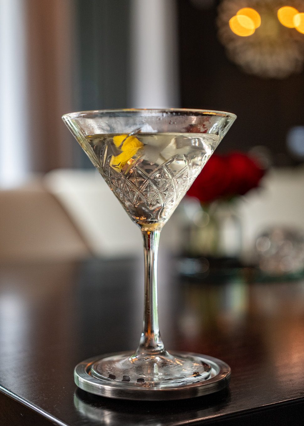 Classic martini served in a crystal-cut glass with a lemon twist, set on a dark bar top with soft ambient lighting and a blurred red rose in the background.