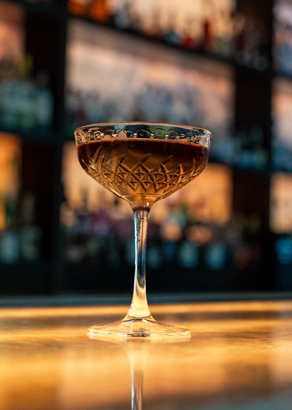 Manhattan cocktail served in an etched coupe glass, filled with a deep amber spirit and set on a softly lit bar top. Backlit shelves of bottles blur in the background, creating a warm, intimate cocktail lounge ambiance.