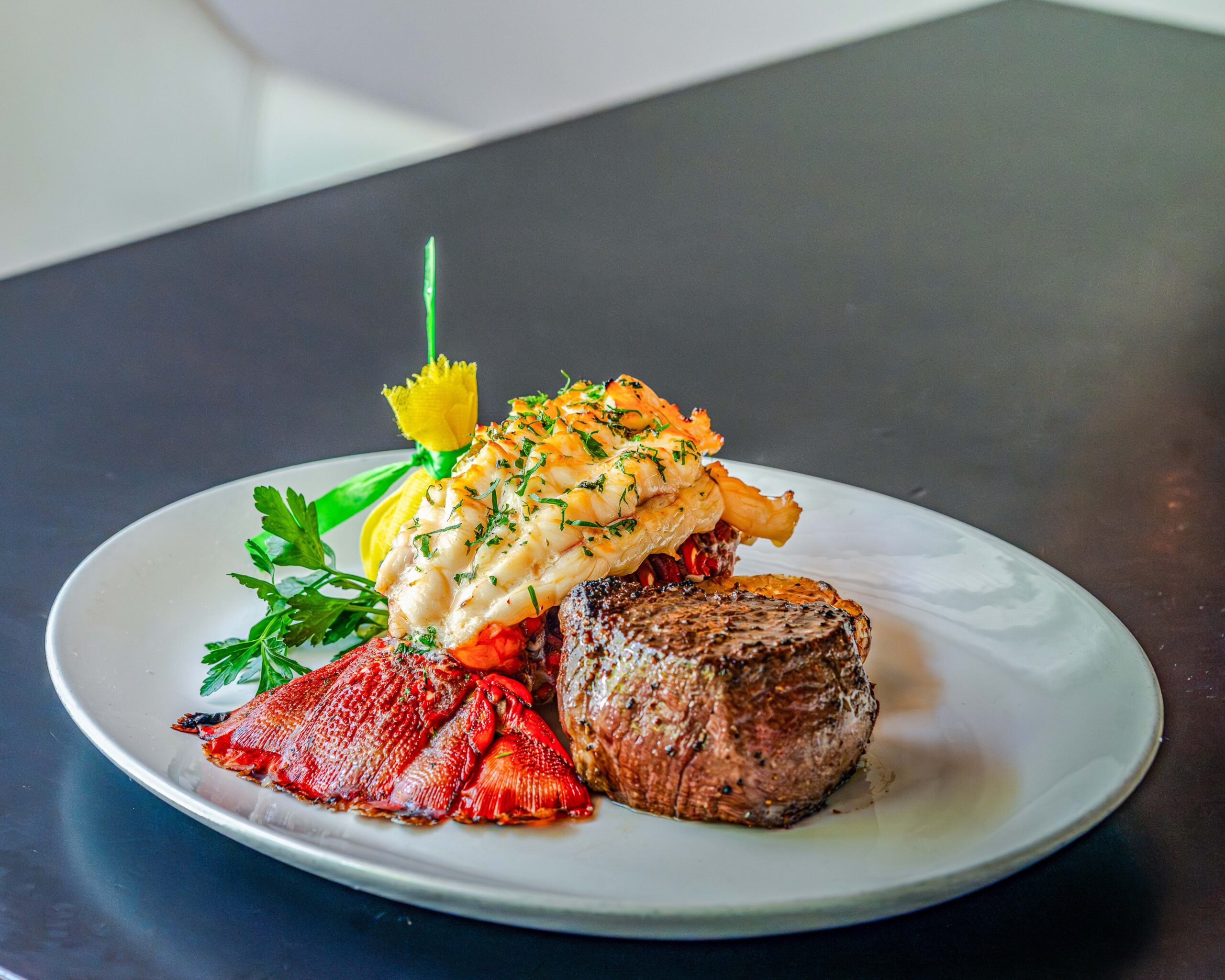 A surf-and-turf entrée featuring a grilled filet mignon topped with a butter-poached lobster tail, garnished with fresh herbs and parsley. The steak and lobster are plated on a white dish against a dark background, highlighting the rich textures and vibrant red lobster shell.