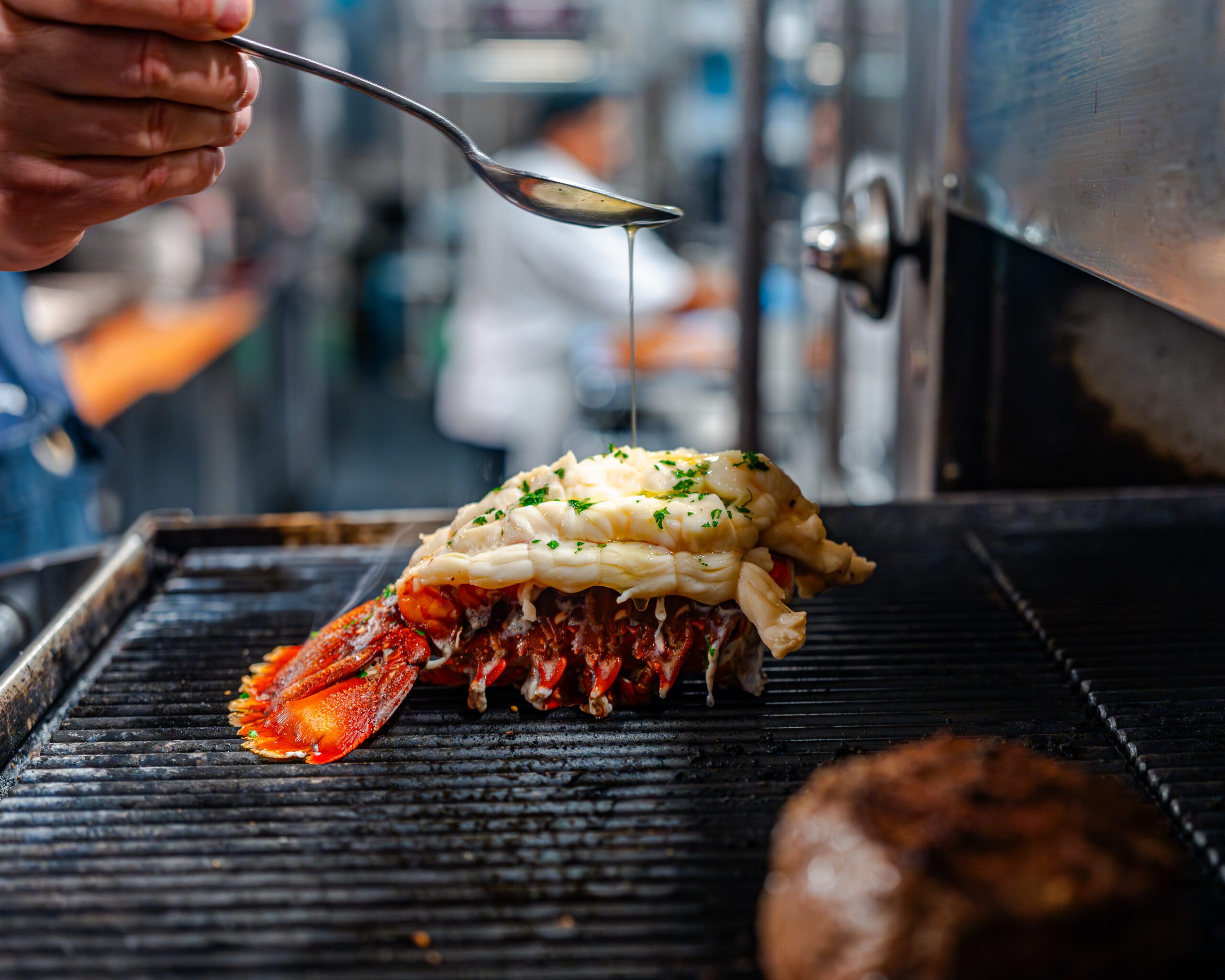 Alt text: Chef drizzling melted butter over a split lobster tail grilling on an open flame, garnished with chopped herbs, with a steak cooking nearby in a professional kitchen setting.