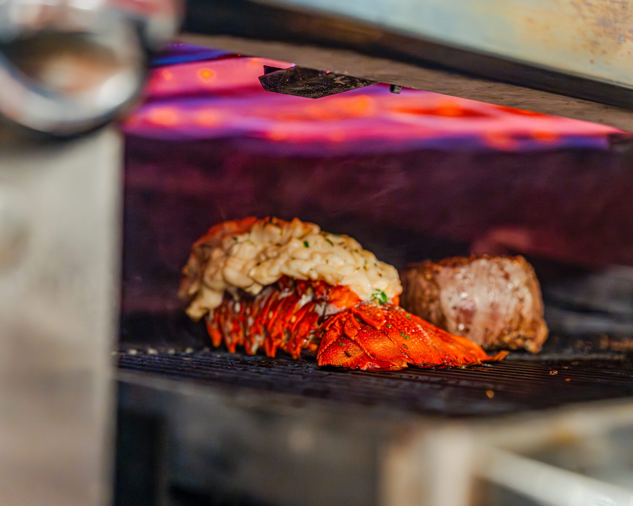 Butter-poached lobster tail and a filet mignon cooking under a high-heat broiler, with flames glowing red in the background. The lobster shell is vibrant red and topped with herbs, highlighting the intense, steakhouse-style preparation.
