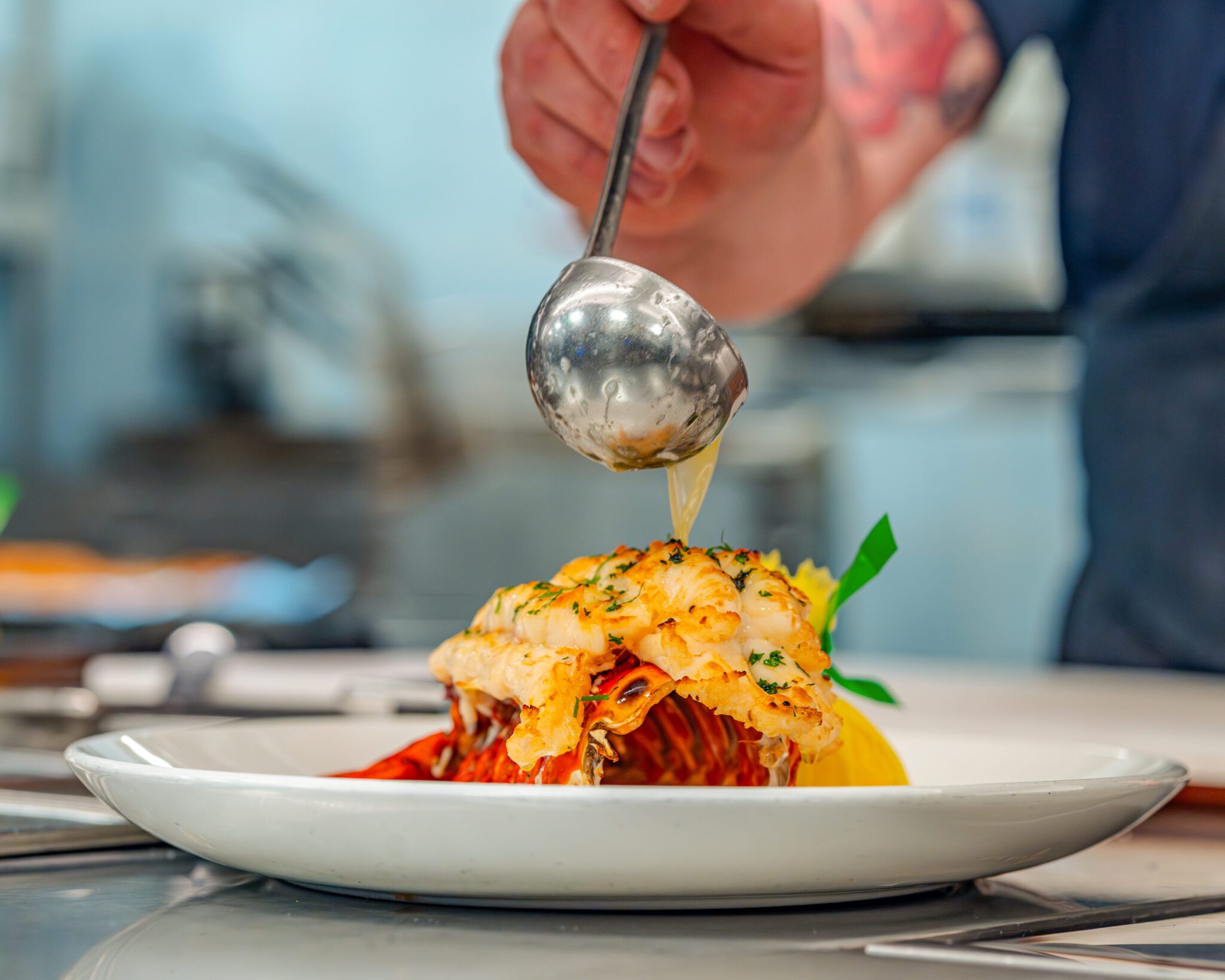 Chef finishing a split lobster tail plated on a white dish, spooning warm melted butter over the tender lobster meat. The bright red shell rests beneath the golden, lightly charred lobster, garnished with fresh herbs and a lemon wedge, with a softly blurred kitchen background.