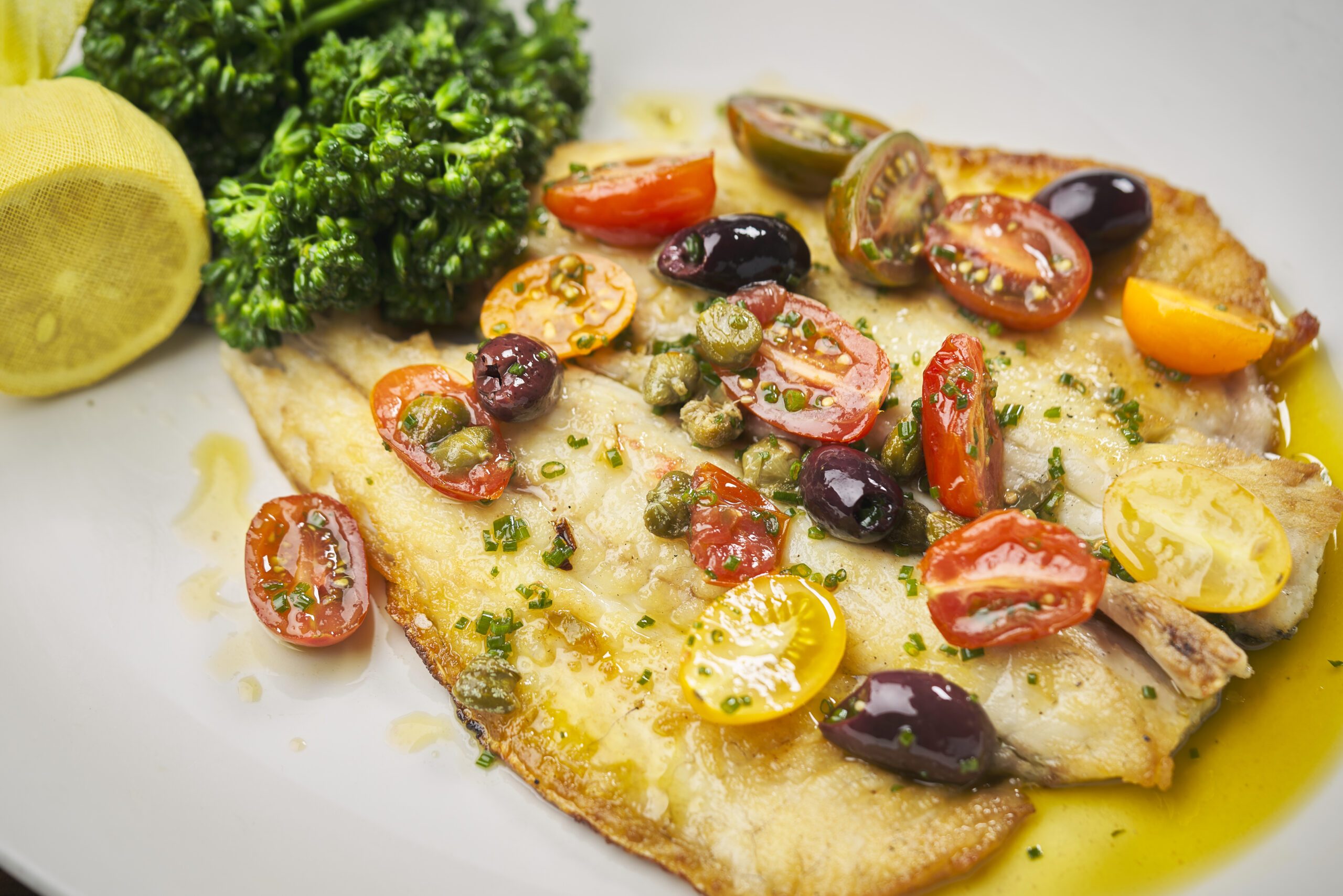 Pan-seared branzino fillet topped with cherry tomatoes, olives, capers, and fresh chives, finished with olive oil. Served with broccolini and a lemon wedge on a white plate.
