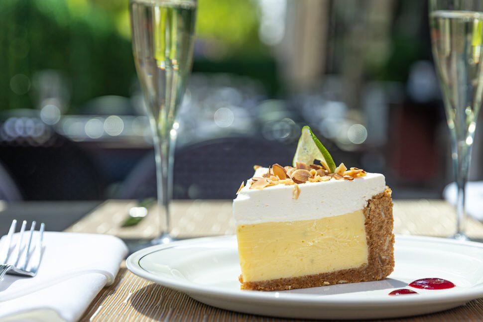 Slice of key lime pie with a graham cracker crust, topped with whipped cream, toasted coconut, and a lime wedge, served on a white plate with raspberry sauce, with two glasses of sparkling wine in the background