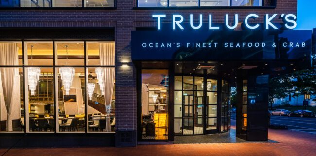 Close-up exterior view of Truluck’s Washington, DC entrance at dusk, featuring illuminated “Ocean’s Finest Seafood & Crab” signage, floor-to-ceiling windows, and a warmly lit dining room visible inside.