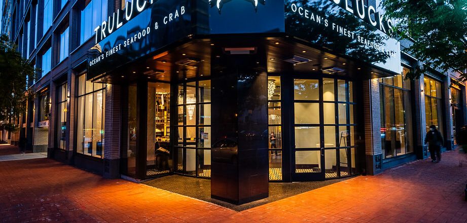 Exterior corner entrance of Truluck’s Ocean’s Finest Seafood & Crab in Washington, DC at dusk, featuring illuminated signage with the crab logo, floor-to-ceiling windows, and warm interior lighting along a brick-lined city sidewalk.