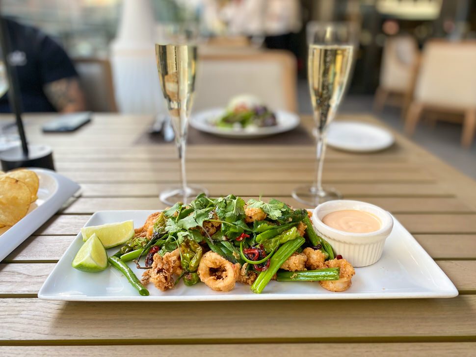 Crispy fried calamari topped with fresh cilantro and sliced red chiles, served with grilled green vegetables, lime wedges, and a creamy dipping sauce on a white rectangular plate, with two glasses of sparkling wine in the background.
