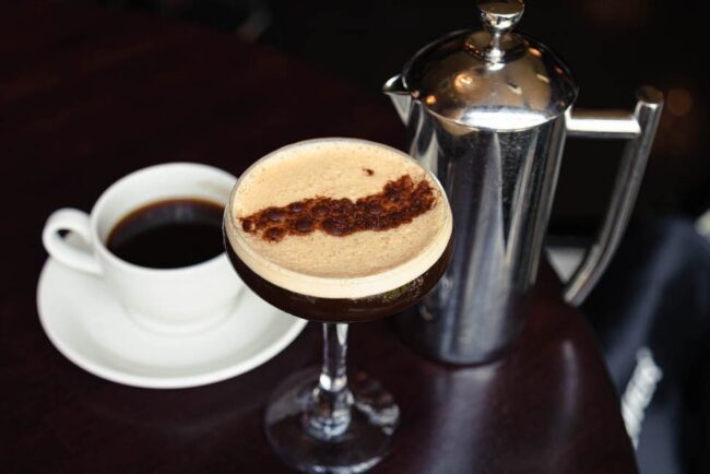 Espresso martini topped with a frothy crema and cocoa dusting, served alongside a cup of coffee and a stainless steel French press on a dark wood table.