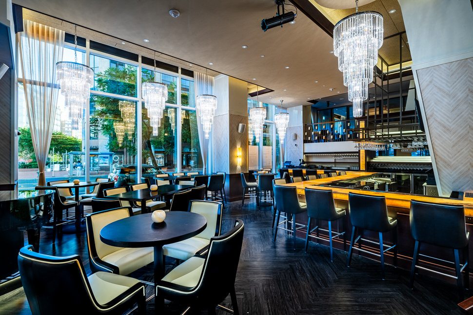 Truluck’s Washington, DC bar featuring a long illuminated bar top with leather bar stools, dramatic crystal chandeliers, floor-to-ceiling windows, and modern lounge seating in an upscale setting.