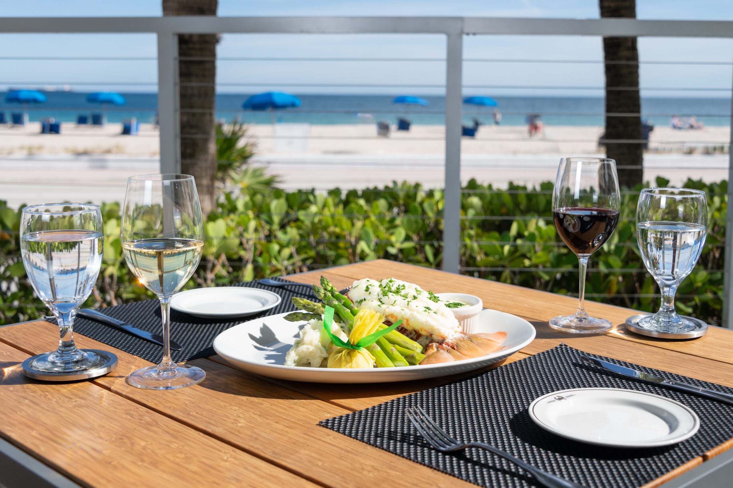 Oceanfront patio dining at Truluck’s Fort Lauderdale featuring a beautifully plated seafood entrée with asparagus and mashed potatoes, paired with red and white wine, set on a sunlit wooden table overlooking the beach and blue Atlantic waters.