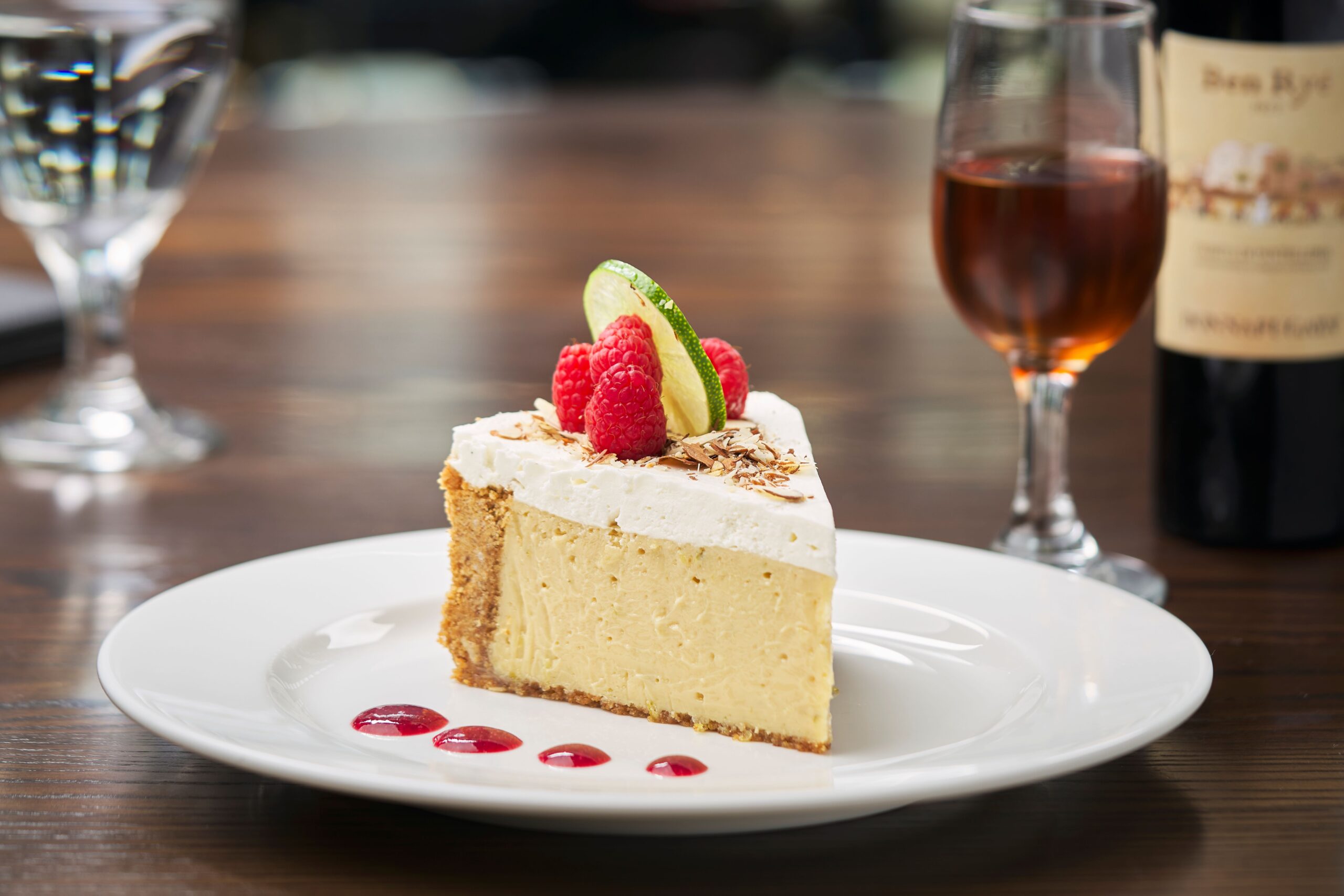 Slice of creamy key lime pie with a graham cracker crust, topped with whipped cream, fresh raspberries, toasted coconut flakes, and a lime wedge, served on a white plate with raspberry sauce dots. A glass of amber dessert wine and wine bottle are softly blurred in the background.