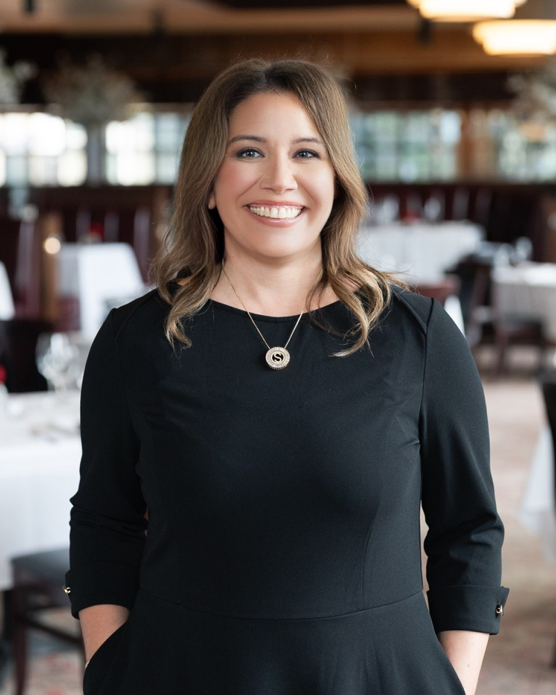 Amber Scheer is standing in Truluck's Woodlands restaurant dining room, wearing a black dress and a gold circular pendant necklace, with white-tablecloth tables softly blurred in the background.