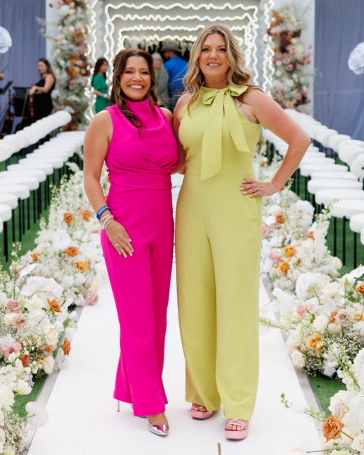 Amber and Lauren are standing together on a floral-lined runway at an outdoor event, smiling toward the camera. One woman wears a bright pink sleeveless jumpsuit and metallic heels, while the other wears a light green jumpsuit with a bow neckline and platform sandals. Rows of white chairs and abundant floral arrangements frame the aisle behind them, with decorative lighting creating an elegant backdrop.