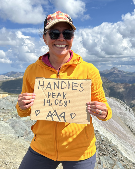 Amber is standing on a mountain summit wearing a bright orange jacket, sunglasses, and a baseball cap, smiling while holding a cardboard sign that reads “Handies Peak 14,058’.” Rocky alpine terrain and distant mountain ranges stretch behind her under a partly cloudy blue sky.