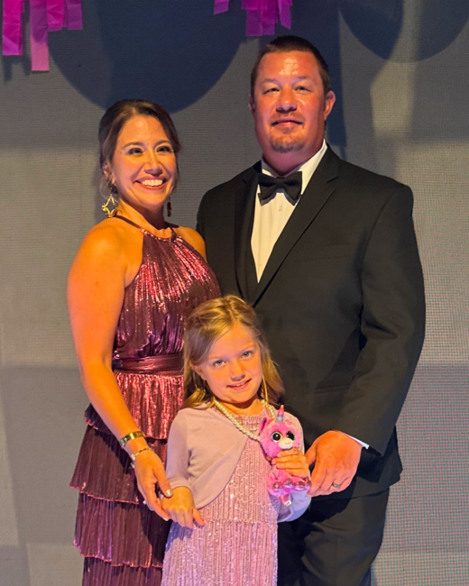 Amber and family posing together at the Hometown Heroes event. Amber is in a burgundy sequined dress, and her husband is in a black tuxedo standing behind a young girl wearing a pink dress and holding a small pink stuffed animal. They smile toward the camera under event lighting with decorative pink accents in the background.