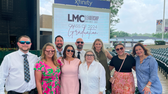 Group of Truluck's team members smiling and posing together the Cynthia Woods Pavilion in front of a large screen reading “LMC Leadership Montgomery County Class of 2024 Graduation.” Rows of empty seats and event staging are visible behind them.