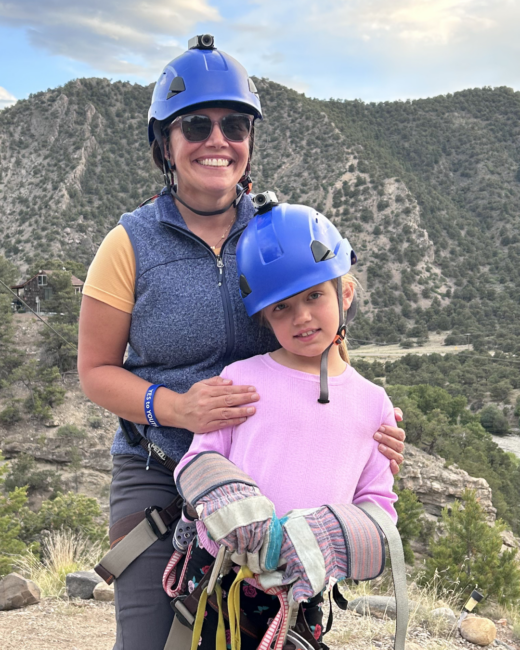 Amber and her daughter wearing blue safety helmets and harnesses stand together outdoors in a mountainous landscape. Amber smiles behind her daughter with her hands on her shoulders while the girl holds climbing gear, with rocky hills and trees in the background.