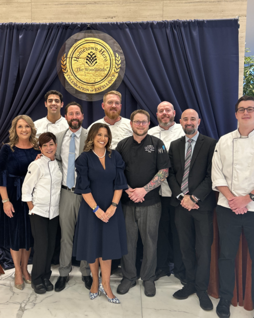 Amber standing with members of the Truluck’s Woodlands team and local chefs at an Interfaith event, posing together in front of a “Hometown Hero – The Woodlands: Celebration of Excellence” backdrop. Several team members wear chef coats while others are dressed in business attire.
