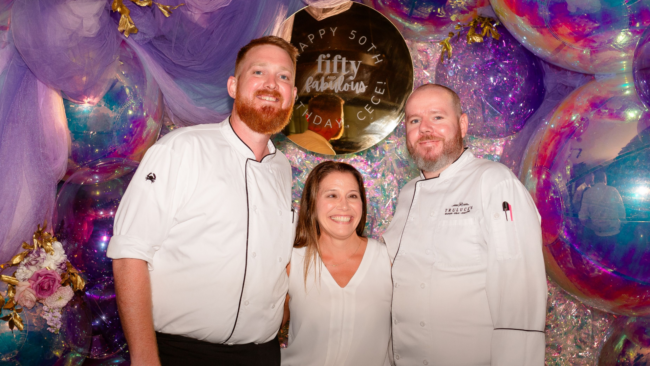 Amber standing with two of her event captains at a celebration, smiling in front of a colorful balloon backdrop and a sign that reads “Happy 50th Birthday Celebration – Fifty and Fabulous.”