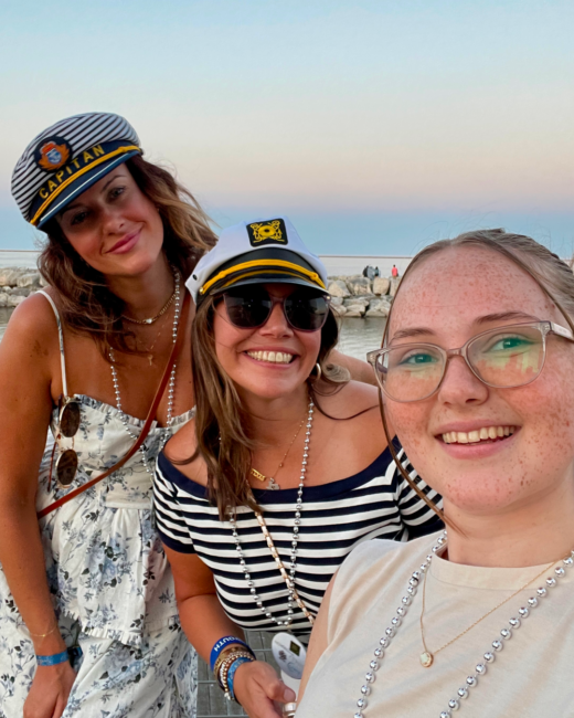 Amber posing for a seaside selfie with Cassandra and Sara at a NACE conference. They smile toward the camera wearing captain-style hats and bead necklaces, with the water and rocky shoreline visible behind them at sunset.