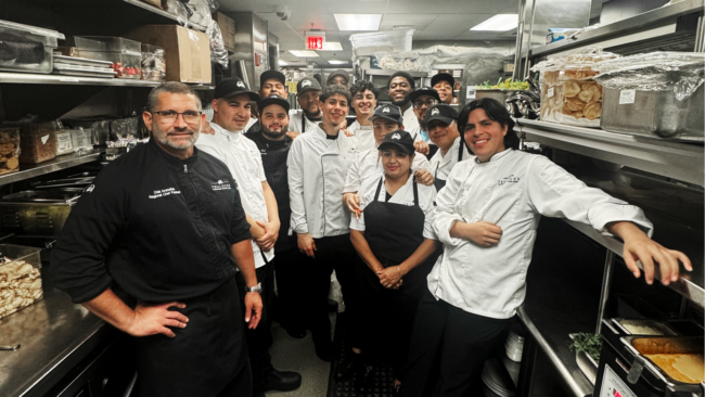 Executive Chef Jonathan Abrevaya stands with the Truluck’s Naples culinary team in the kitchen, posing together in chef coats and caps between stainless steel prep stations and shelves, showcasing the camaraderie and teamwork behind the restaurant’s cuisine.