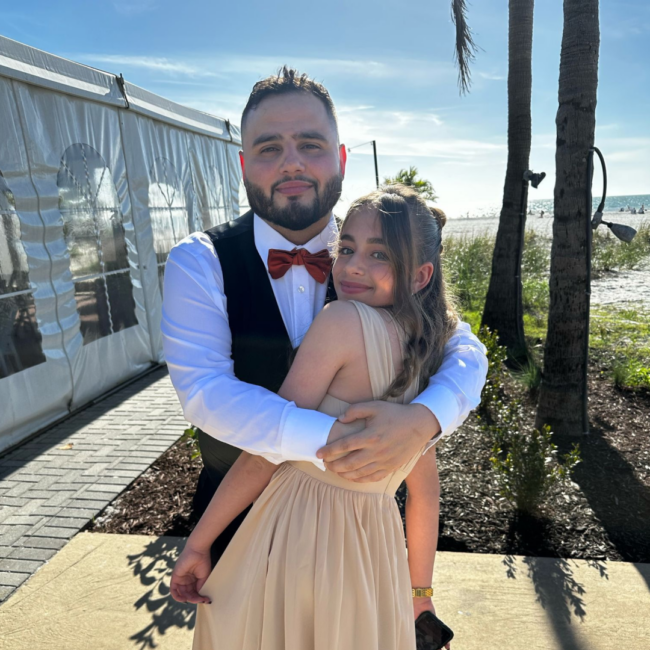 Chef Jonathan Abrevaya embraces a family member outdoors by the beach, dressed in formal attire with a red bow tie and black vest, as palm trees, a white event tent, and the sunlit shoreline create a warm, celebratory backdrop.