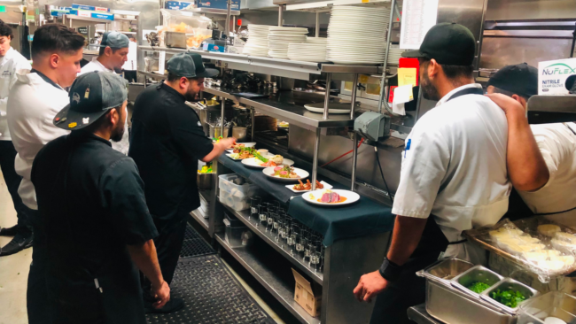 Chef Jonathan Abrevaya leads the Truluck’s Naples culinary team during service, as plated entrées line the pass and chefs in uniforms and caps focus on final garnishes in the bustling kitchen.