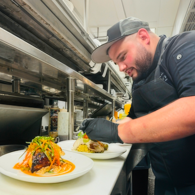 Executive Chef Jonathan Abrevaya carefully plates a dish in the Truluck’s kitchen, wearing a black chef coat, apron, cap, and gloves as he garnishes an entrée with vibrant vegetables and sauce at the pass.