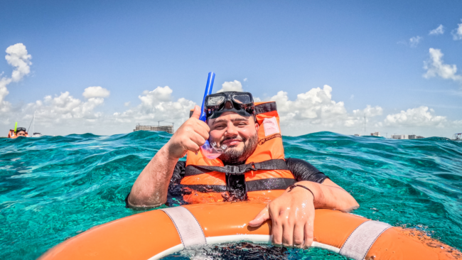 Chef Jonathan Abrevaya floats in clear turquoise water wearing a snorkel mask and orange life vest, smiling and giving a thumbs-up while holding onto a flotation ring under a bright blue sky with scattered clouds and distant shoreline buildings in the background.