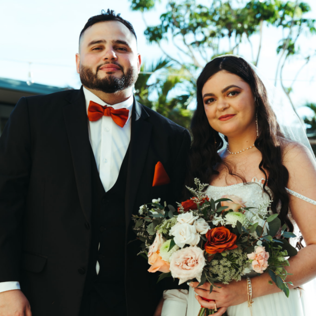 Chef Jonathan Abrevaya and his bride pose outdoors on their wedding day, standing close together in formal attire. He wears a black tuxedo with a red bow tie and matching pocket square, while she wears an off-the-shoulder white gown and holds a bouquet of ivory, blush, and red roses with greenery. Palm trees and soft natural light create a warm, elegant backdrop.