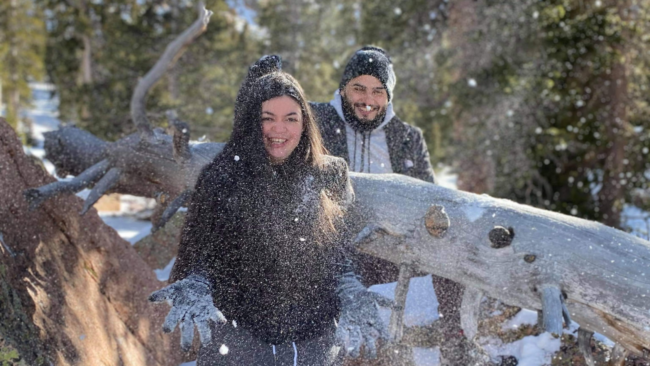 Chef Jonathan Abrevaya and his wife enjoy a snowy day outdoors, bundled in winter coats and hats as they smile and toss snow near fallen tree branches in a wooded setting.