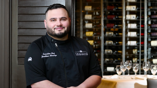 Executive Chef Jonathan Abrevaya of Truluck’s Naples stands in the dining room wearing a black chef coat embroidered with his name and the Truluck’s logo, smiling confidently in front of a glass wine wall and elegantly set tables at Truluck's Naples.