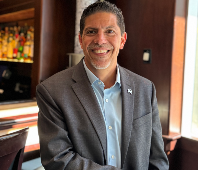 Mickey Gonzalez, General Manager of Truluck’s Miami, smiling in a gray blazer and light blue shirt inside an elegant restaurant bar with warm wood accents and softly lit bottles in the background.
