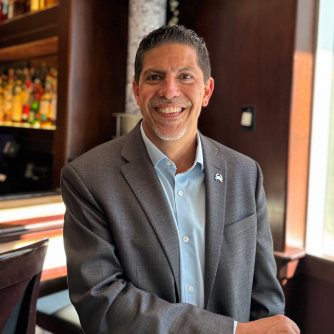 Mickey Gonzalez, General Manager of Truluck’s Miami, smiling in a gray blazer and light blue shirt inside an elegant restaurant bar with warm wood accents and softly lit bottles in the background.