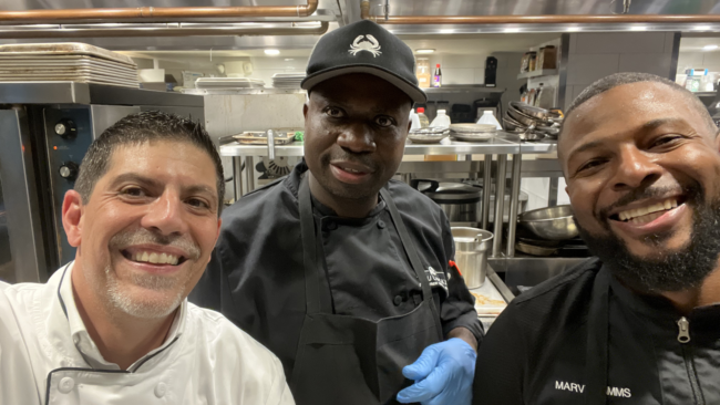 Mickey Gonzalez, General Manager of Truluck’s Miami, smiling with culinary team members in a professional kitchen, surrounded by stainless steel equipment and prep stations.