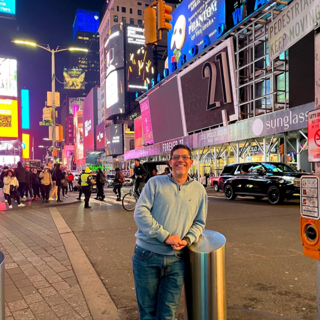Mickey Gonzalez, General Manager of Truluck’s Miami, standing in Times Square at night, smiling amid bright city lights, digital billboards, and bustling street activity.