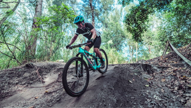 Mickey Gonzalez, General Manager of Truluck’s Miami, mountain biking on a wooded trail, wearing a helmet and cycling gear while navigating a dirt path surrounded by lush greenery.