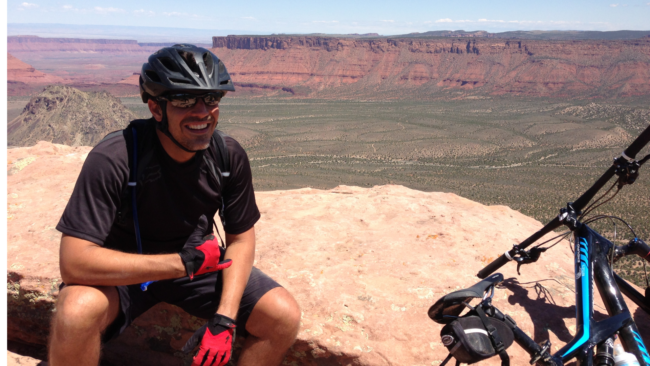 Mickey Gonzalez, General Manager of Truluck’s Miami, mountain biking at a scenic desert overlook, wearing a helmet and gear with expansive canyon views in the background.