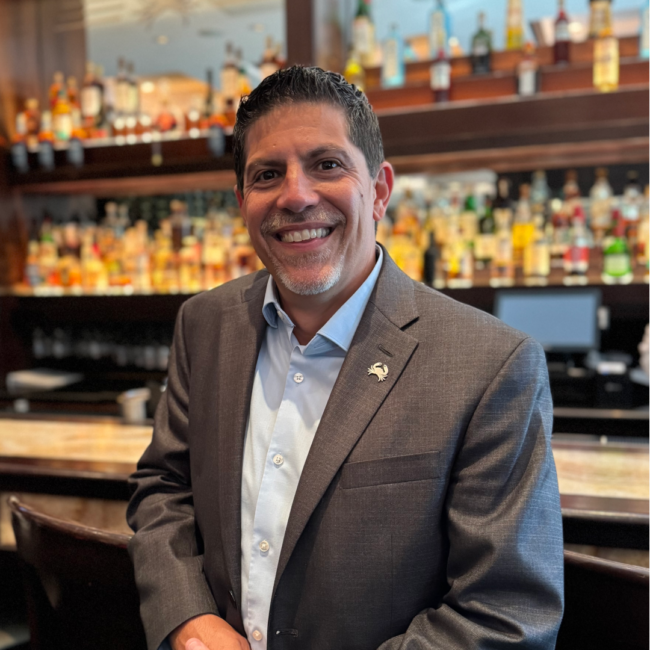 Mickey Gonzalez, General Manager of Truluck’s Miami, smiling in a gray blazer and light blue shirt while standing at the restaurant bar, with a backdrop of premium spirits and warm, elegant interiors.
