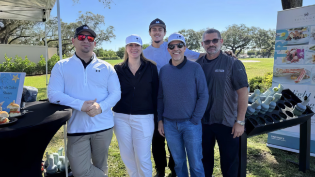 Mickey Gonzalez, General Manager of Truluck’s Miami, posing with colleagues at an outdoor event on a sunny day, standing on a lawn beside a Truluck’s display featuring seafood offerings and branded materials.
