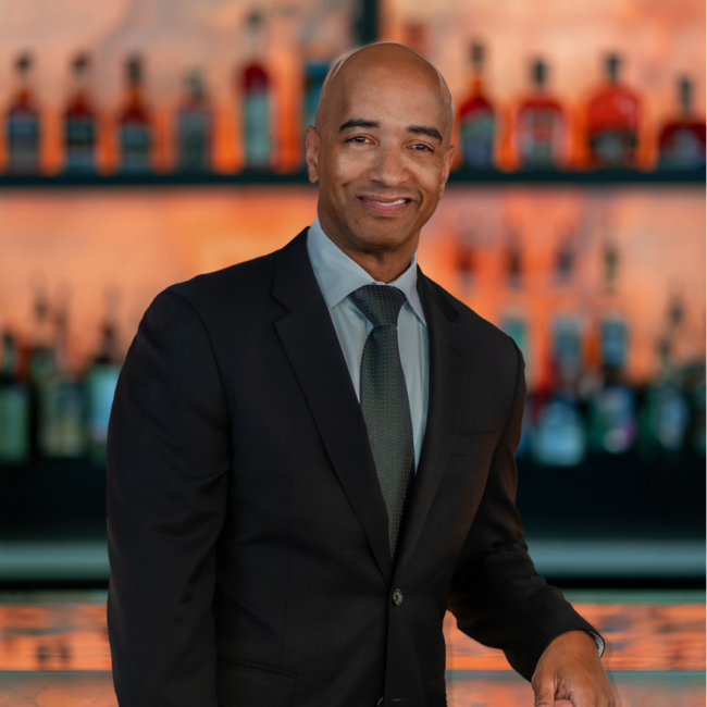 Anthony Ray, General Manager of Truluck’s Washington, D.C., stands in a tailored dark suit and tie, smiling warmly in front of an elegant, softly lit bar lined with premium spirits.
