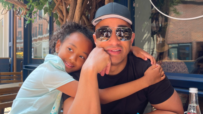 Anthony Ray sits outdoors at a restaurant table, wearing a black shirt, baseball cap, and reflective sunglasses, while a young child with curly hair leans in affectionately with an arm around his shoulders.