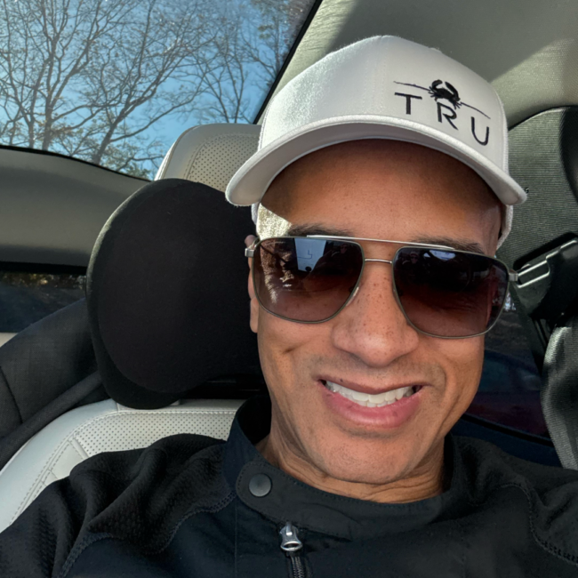 Anthony Ray smiles in a close-up selfie taken inside a car, wearing a white TRU-branded baseball cap, dark sunglasses, and a black zip-up top, with sunlight and tree branches visible through the window behind him.