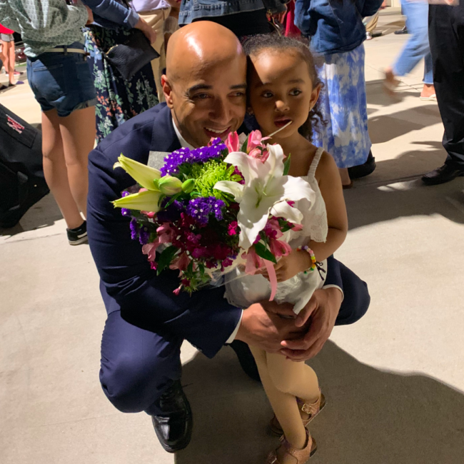 Anthony Ray kneels beside a young child at what appears to be a celebration or event, both smiling as they hold a vibrant bouquet of flowers featuring lilies and colorful blooms, surrounded by a lively crowd in the background.