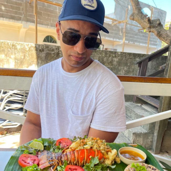 Anthony Ray sits outdoors at a casual dining spot, wearing a white T-shirt, sunglasses, and a navy baseball cap, looking down at a plated lobster dish served with fresh vegetables and sauce on a large green leaf.